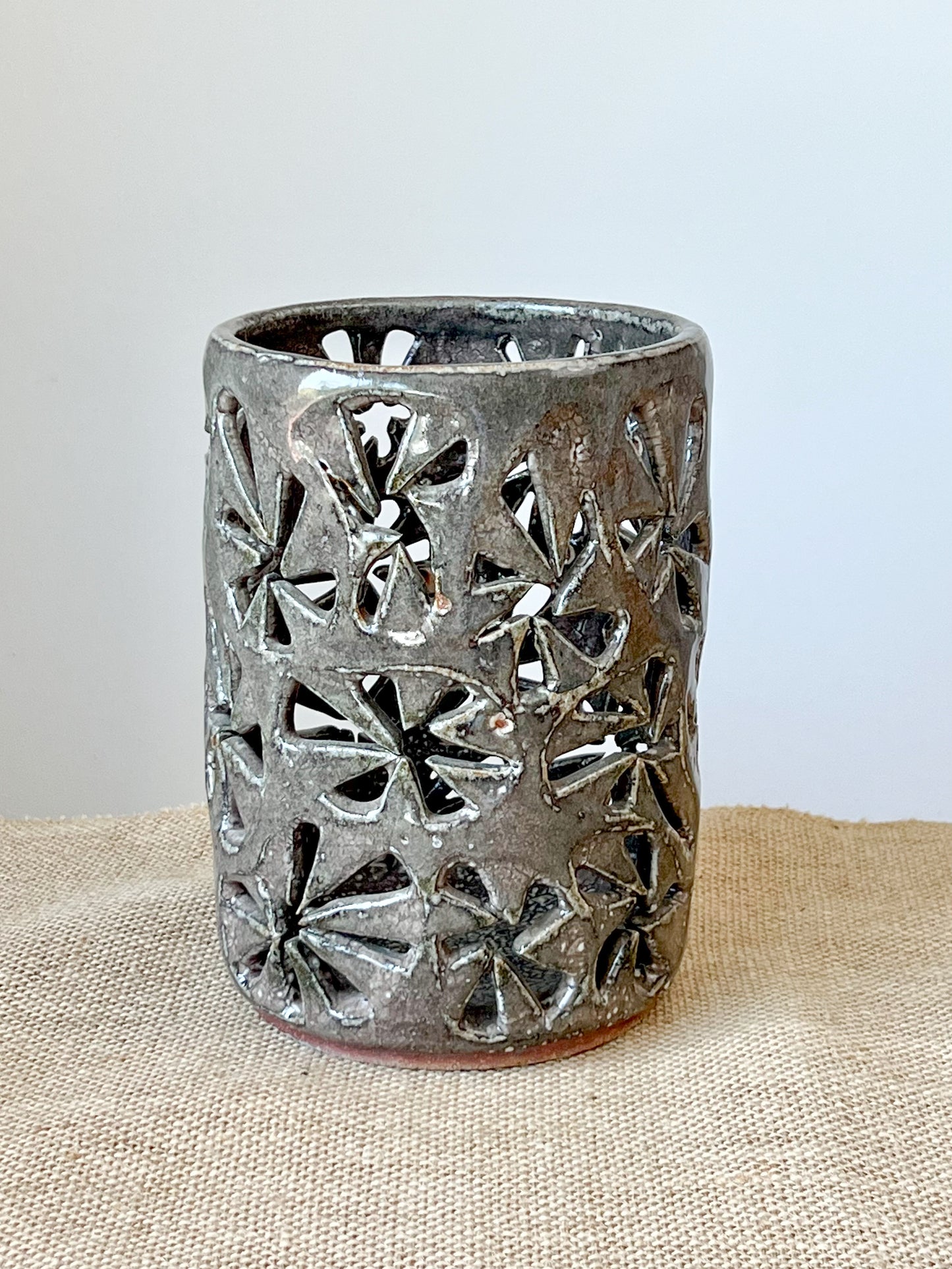 Wheel thrown using stoneware clay. Hand carved with pinwheel pattern to let light through. A warm gray glaze covers the piece. Simply add a candle to fairy lights.
