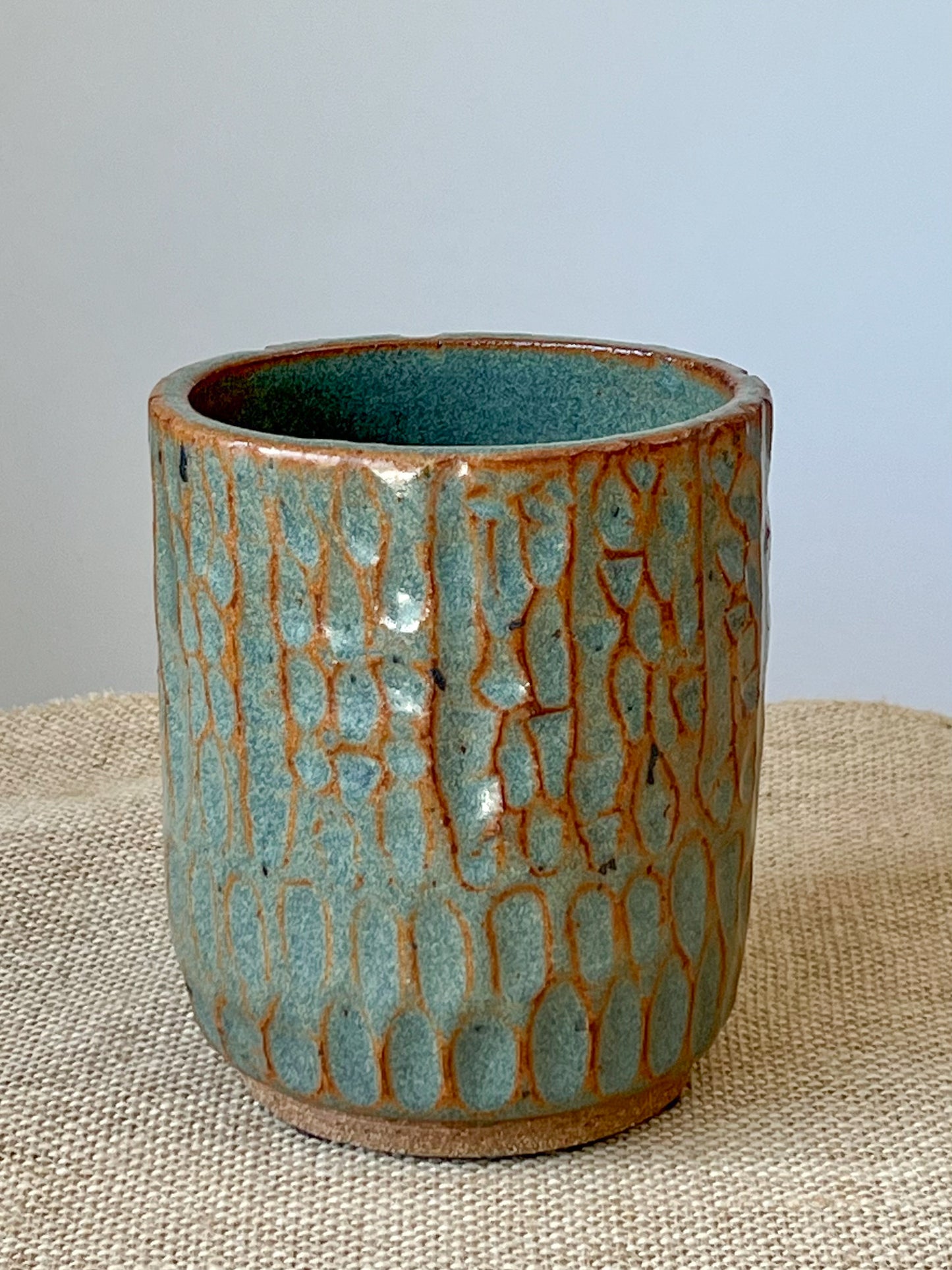 Wheel thrown cylinder using brown stoneware clay. Top rim is altered to an oval. Hand carved texture is highlighted with the blue green glaze.
